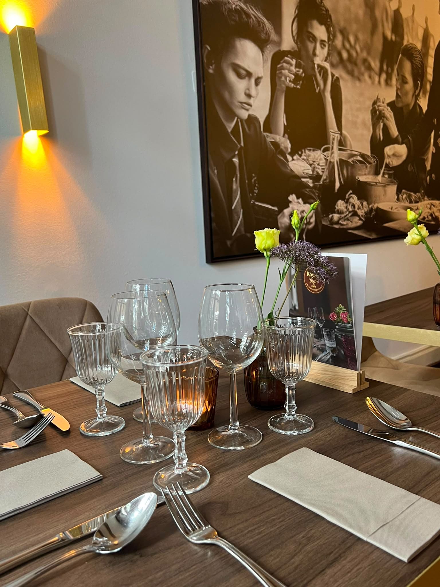 Gedeckter Tisch mit Weingläsern und Blumen vor einer leuchtenden Messing-Wandleuchte