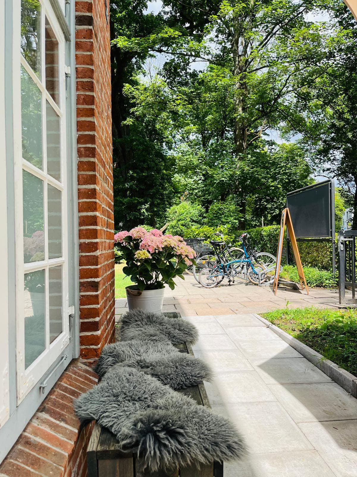 Sonnige Bank mit Lammfellen an der Backsteinwand, Hortensie im Topf, dahinter Garten und hohe Bäume