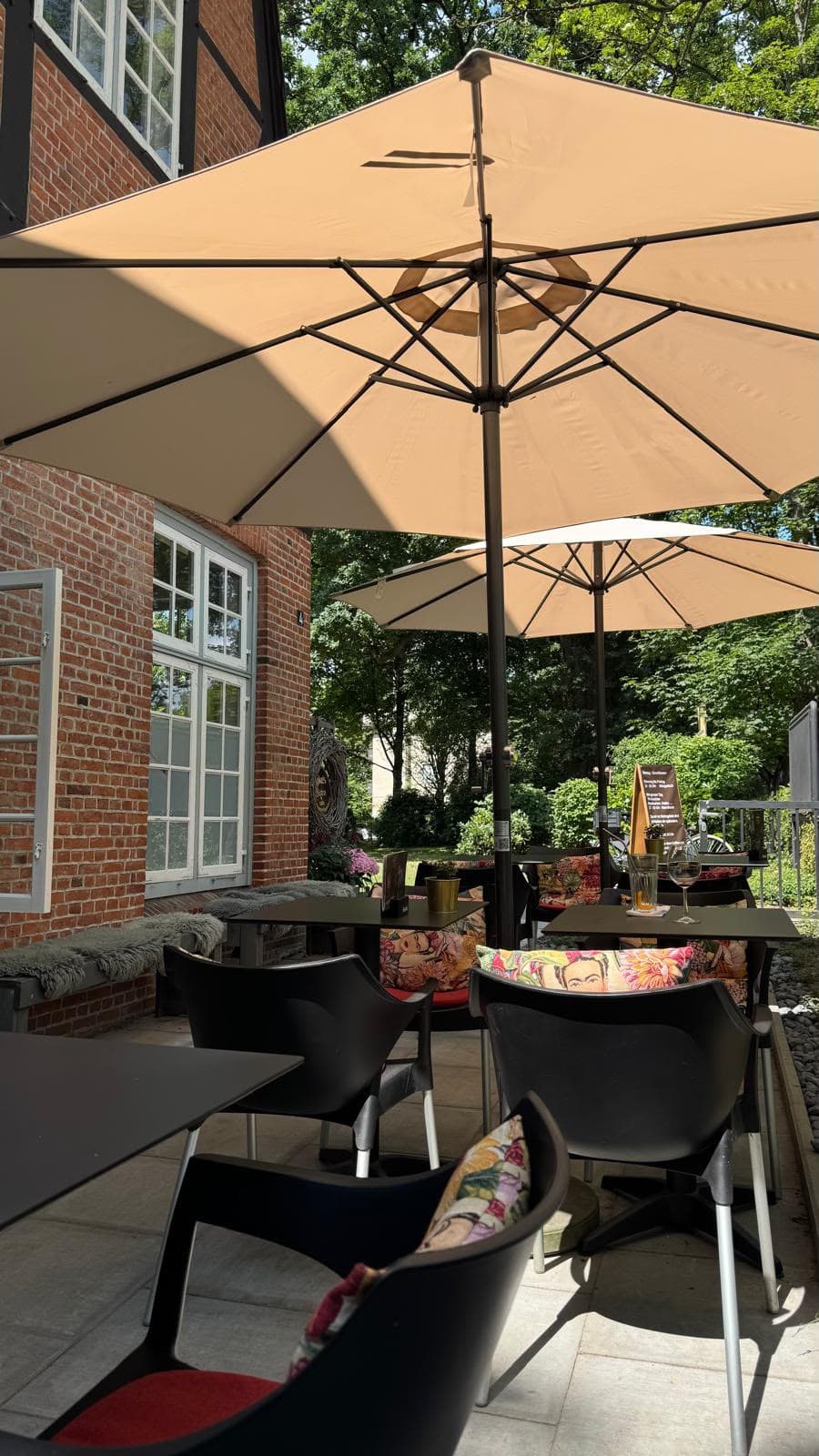 Terrasse unter hellen Sonnenschirmen, schwarze Stühle mit bunten Kissen, Backsteinfassade und Bäume